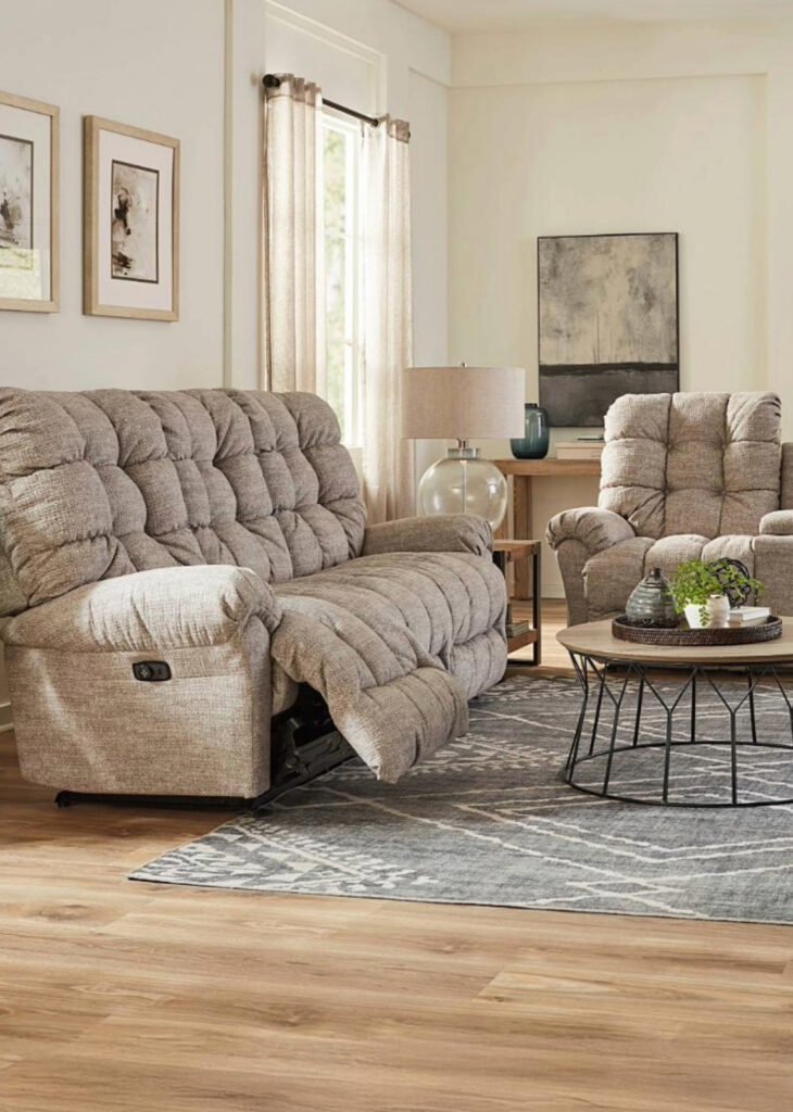  A cozy living room featuring a plush, tufted reclining sofa and matching recliner in a soft, neutral fabric. The sofa is partially reclined, showcasing its comfort and functionality. A round coffee table with a wireframe base sits atop a patterned area rug, decorated with a tray and greenery. In the background, a wooden side table with a glass lamp and decorative accents adds warmth to the space. Large windows with sheer curtains allow natural light to brighten the neutral-toned room.