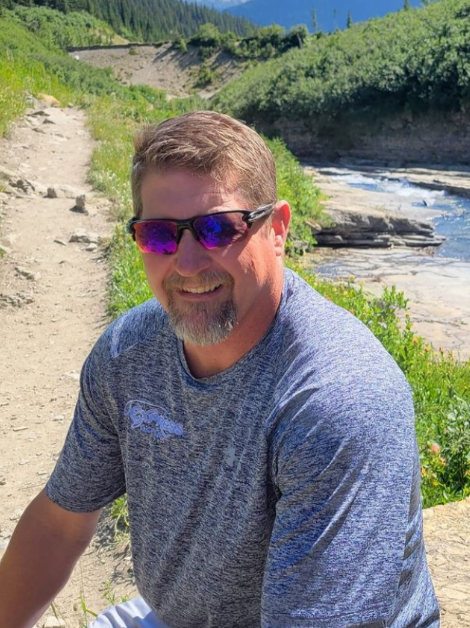 Meyers family member enjoying the outdoors on a hiking trail near a scenic river, wearing sunglasses and a blue shirt.